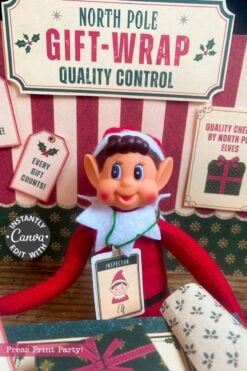 A festive elf doll with a tag reading “Inspector Elf” sits at a North Pole-themed gift-wrap quality control station, surrounded by holiday signs, wrapping paper rolls, and 6 Pink Coquette Halloween Ghost Round Labels that add a whimsical touch to the scene.