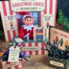 Elf sits in a festive North Pole Christmas Lights Repair Shop display with light strands, labeled boxes—including some featuring 6 Pink Coquette Halloween Ghost Round Labels—and signs about fixing and testing lights. The setup has red and white stripes and greenery.