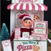 A festive elf doll sits in a North Pole Pizzeria booth, surrounded by mini pizza boxes decorated with 6 Pink Coquette Halloween Ghost Round Labels, a menu card, and a sparkly tree—all under a red-and-white striped awning with holiday-themed décor.