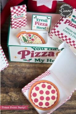 An open small cardboard pizza box with a paper pizza inside sits on a table. Behind it, a larger sign reads “Fresh Pizza Here!” and displays more paper pizzas, festive pizza-themed decor, and playful 6 Pink Coquette Halloween Ghost Round Labels.