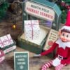 An Elf doll sits next to a festive North Pole package scanner display decorated with a wrapped gift box, a “Parcel Sorting” sign, and colorful string lights in the background. A green paper panel reads “Press Print Party!” while 6 Pink Coquette Halloween Ghost Round Labels add a charming seasonal touch to the scene.