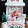 A festive North Pole Sweet Shop display features an elf doll, candy boxes, a mini Christmas tree, and a menu with holiday treats. A striped awning and candy-themed signs create a playful, holiday atmosphere, accented by 6 Pink Coquette Halloween Ghost Round Labels for an extra touch of charm.