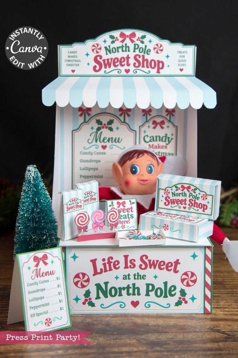 A festive North Pole Sweet Shop display features an elf doll, candy boxes, a mini Christmas tree, and a menu with holiday treats. A striped awning and candy-themed signs create a playful, holiday atmosphere, accented by 6 Pink Coquette Halloween Ghost Round Labels for an extra touch of charm.