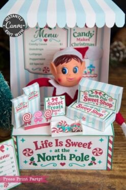 A festive elf doll sits behind a “Life Is Sweet at the North Pole” candy stand adorned with mini boxes, sweets, and a holiday-themed menu in the background, featuring 6 Pink Coquette Halloween Ghost Round Labels that add an extra touch of whimsy to the scene.