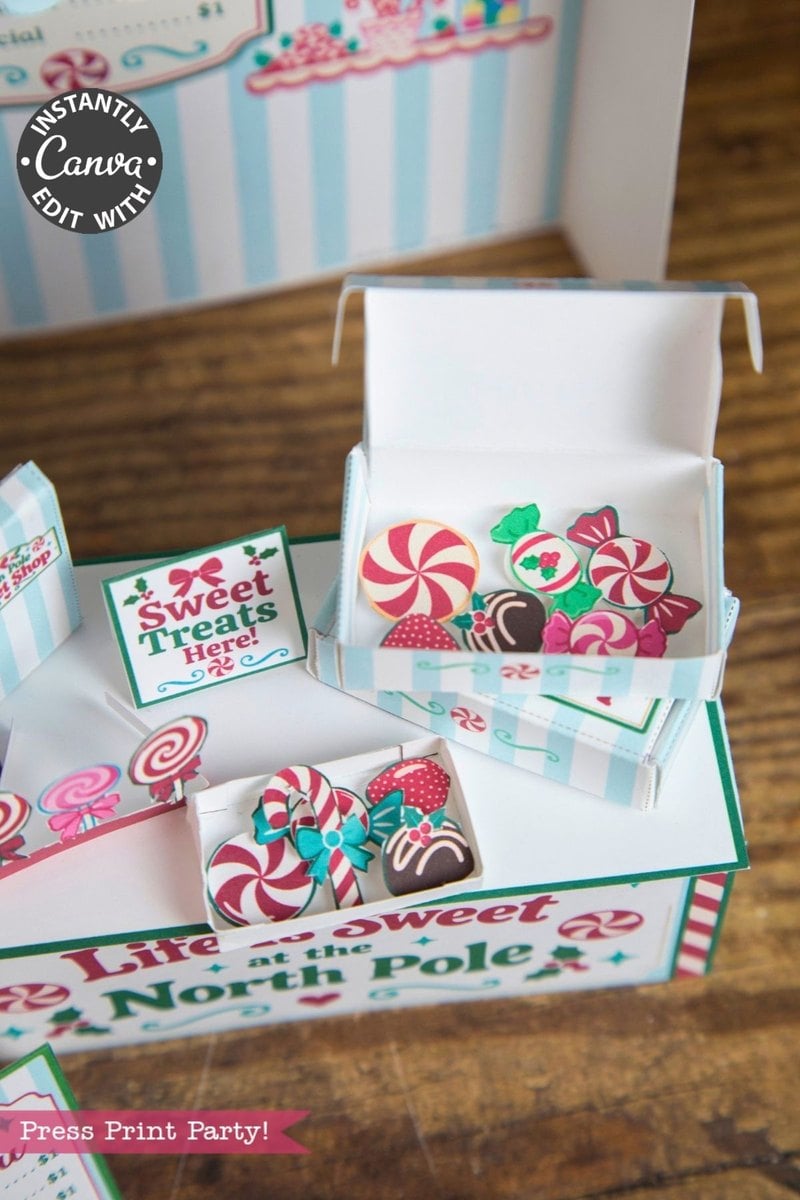 Open white boxes filled with illustrated candy cutouts, including peppermint swirls and chocolates, rest on a wooden surface. Festive signs and “Life is Sweet at the North Pole” text decorate the scene, while 6 Pink Coquette Halloween Ghost Round Labels add a charming touch to this whimsical display.