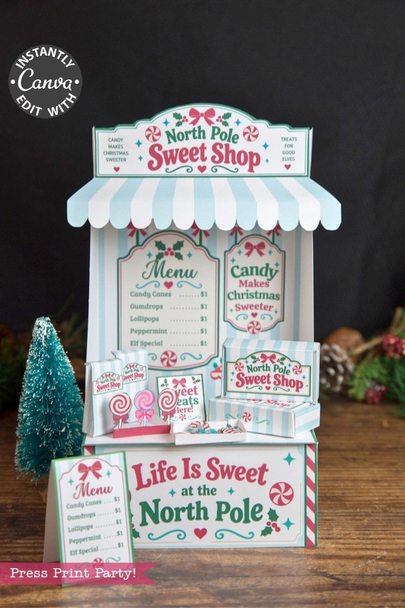 A miniature “North Pole Sweet Shop” display with a striped awning, holiday candy menus, festive signs, and lollipops—each decorated with 6 Pink Coquette Halloween Ghost Round Labels—set on a table surrounded by small pine trees and Christmas decorations in the background.