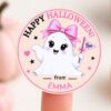 A round sticker from the 6 Pink Coquette Halloween Ghost Round Labels set features a cute, smiling ghost holding a “Boo” flag, wearing a pink bow. The label displays editable text reading “Happy Halloween! from EMMA.” The sticker is pictured on a finger with pumpkins blurred in the background.