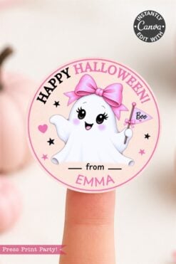 A round sticker from the 6 Pink Coquette Halloween Ghost Round Labels set features a cute, smiling ghost holding a “Boo” flag, wearing a pink bow. The label displays editable text reading “Happy Halloween! from EMMA.” The sticker is pictured on a finger with pumpkins blurred in the background.