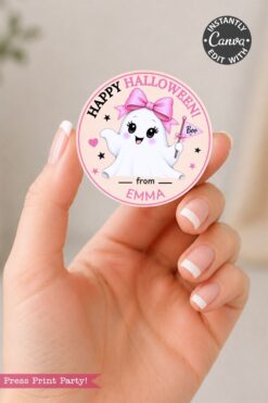 A hand holds one of the 6 Pink Coquette Halloween Ghost Round Labels—a round pink sticker featuring a cute ghost with a pink bow and magic wand. The label is ideal for Pink Coquette Halloween projects, displaying the text “Happy Halloween from Emma,” along with the words “Boo” and “Instantly Canva edit with.”.