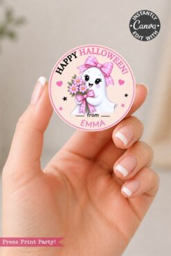 A hand holds one of the 6 Pink Coquette Halloween Ghost Round Labels, featuring a charming ghost with a pink bow and flowers. The pastel-colored sticker displays the cheerful message, “Happy Halloween from Emma,” making it perfect for your festive labeling needs.