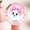 A round pink sticker from the 6 Pink Coquette Halloween Ghost Round Labels collection showcases a cute smiling ghost wearing a pink polka-dot bow with the text “Happy Halloween! from EMMA.” The sticker is displayed on a fingertip, with softly blurred pumpkins in the background.