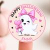 A round label from the 6 Pink Coquette Halloween Ghost Round Labels set features a cute ghost holding a heart-shaped wand, both adorned with pink bows and “Happy Halloween” written at the top. “From Emma” is inscribed at the bottom, and the tag is delicately balanced on someone’s fingertip, reflecting the sweet coquette style of these Halloween labels.