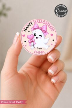 A hand holds a round sticker from the 6 Pink Coquette Halloween Ghost Round Labels set, featuring a cute ghost with a pink bow, holding a heart wand, and the text “Happy Halloween! from EMMA” on a pink background. The Canva logo and “Press Print Party!” text are also visible.