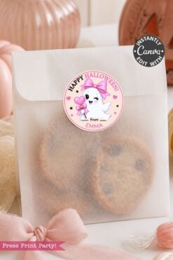 A frosted bag of cookies is sealed with one of the 6 Pink Coquette Halloween Ghost Round Labels, which features a cute ghost, a bow, and the message “Happy Halloween from Emma.” The scene is decorated with pink accents and pumpkins in the background.