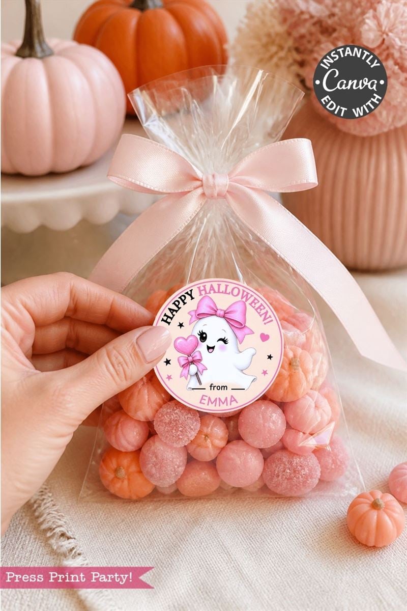 A hand holds a clear bag of pink candies tied with a pink ribbon, featuring one of the 6 Pink Coquette Halloween Ghost Round Labels, which display a cute ghost, the text “Happy Halloween,” and space for a name. Pink pumpkins and decorations are in the background.