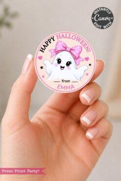 A hand holds a round sticker featuring a cute ghost with a pink bow, the words “Happy Halloween from Emma,” and pink accents. The design promotes easy editing with Canva.