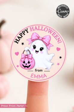 A finger holds a round, pink Halloween sticker featuring a cute ghost with a pink bow, holding a pink jack-o’-lantern. Text reads “Happy Halloween! from EMMA.” Soft pink pumpkins are blurred in the background.