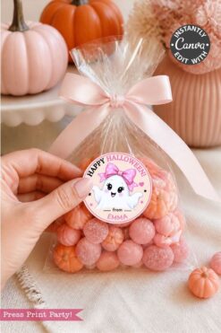 A hand holds a clear bag of pink pumpkin candies with a “Happy Halloween” ghost sticker. A pink bow ties the bag, surrounded by pastel pumpkins on a table.