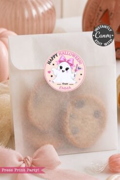 A translucent bag with chocolate chip cookies inside, sealed with a “Happy Halloween” sticker featuring a cute ghost. Pink decorations and pumpkins are in the background.