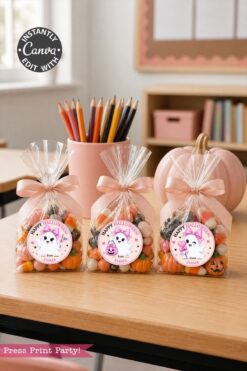 Three clear bags of Halloween candy with personalized tags sit on a classroom desk. Colored pencils, books, and a pink pumpkin are in the background.