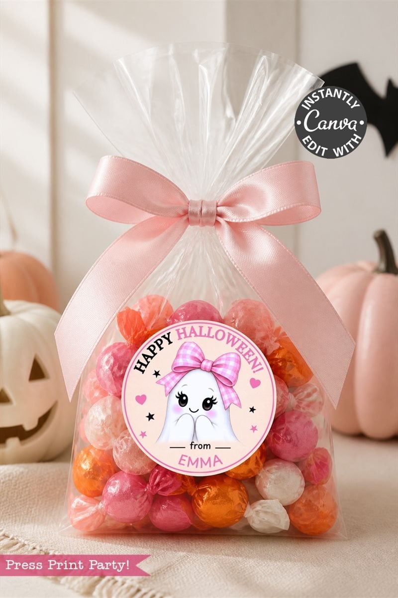A clear bag filled with pink, orange, and white candies is tied with a pink ribbon and adorned with one of the 6 Pink Coquette Halloween Ghost Round Labels reading “Happy Halloween from Emma”; the bag sits surrounded by pumpkins.
