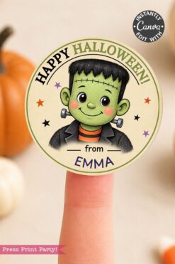 A finger holds a round Halloween sticker featuring a cute cartoon Frankenstein and text reading “Happy Halloween! from EMMA.” Pumpkins are blurred in the background.