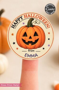 A finger holds a round Halloween sticker featuring a smiling jack-o’-lantern and the text “Happy Halloween! from Emma.” A pumpkin and blurred objects are in the background.