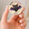 A hand holds a round, pink Coquette Ghost Treat Bag Halloween sticker featuring a cute bat with “Happy Halloween from Emma” written on it, along with stars and “Instantly Edit with Canva” in the corner.
