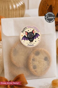 A clear bag filled with chocolate chip cookies is sealed with a Pink Coquette Ghost Treat Bag Halloween Sticker, which features a cute bat and the text “Happy Halloween from Emma.” In the background, there is a pumpkin and ribbon.