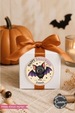 A white gift box with an orange ribbon sits on a table. Adorning the box is a round Pink Coquette Ghost Treat Bag Halloween Sticker featuring a cute bat and the words “Happy Halloween from Emma.” In the background, there’s a pumpkin, candle, and beads.