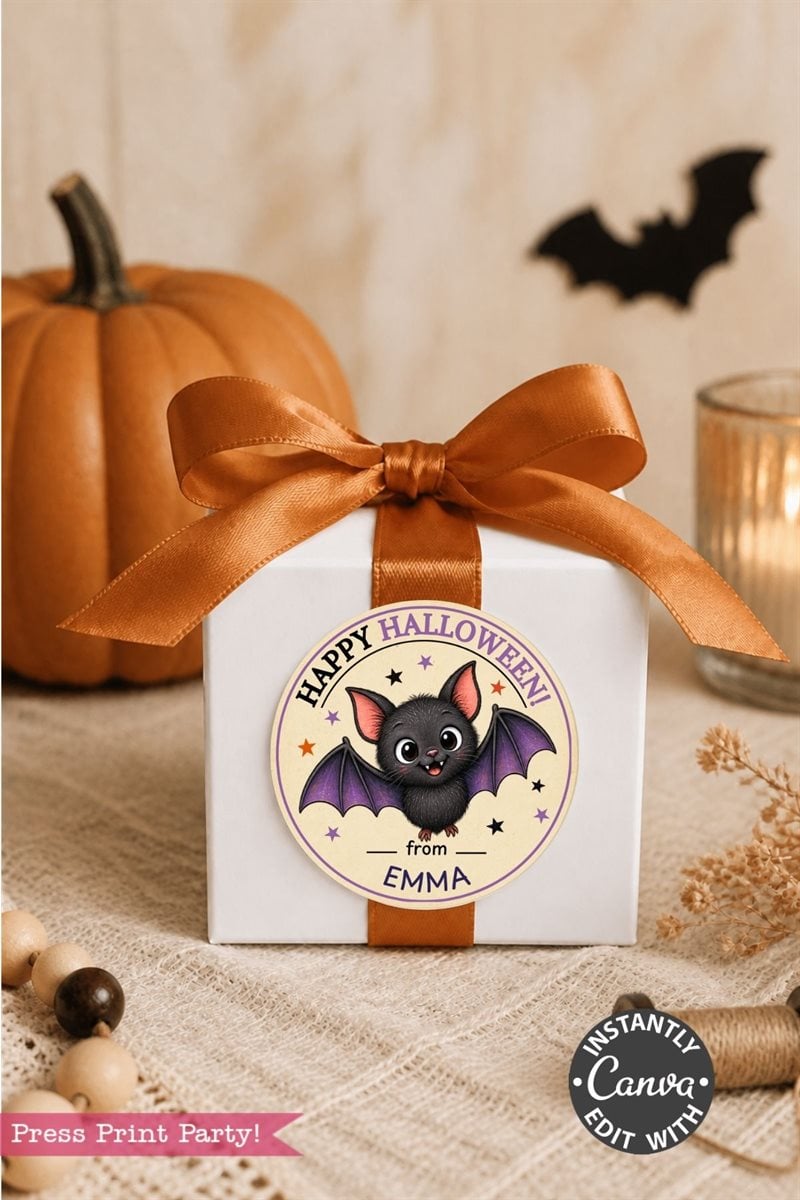 A white gift box with an orange ribbon sits on a table. Adorning the box is a round Pink Coquette Ghost Treat Bag Halloween Sticker featuring a cute bat and the words “Happy Halloween from Emma.” In the background, there’s a pumpkin, candle, and beads.