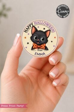 A hand holds a round, pink Coquette Ghost Treat Bag Halloween sticker featuring a cute black cat with an orange bow tie and the message “Happy Halloween from Emma,” surrounded by stars.