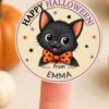 A finger holds a round, pink Pink Coquette Ghost Treat Bag Halloween Sticker featuring a cute black cat in an orange polka dot bow tie, surrounded by stars and the text “Happy Halloween from Emma.” Pumpkins are blurred in the background.
