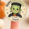 A Pink Coquette Ghost Treat Bag Halloween Sticker featuring a cute cartoon Frankenstein and the text “Happy Halloween! from EMMA” is held on a fingertip, adding an adorable touch to your celebrations with blurred pumpkins in the background.