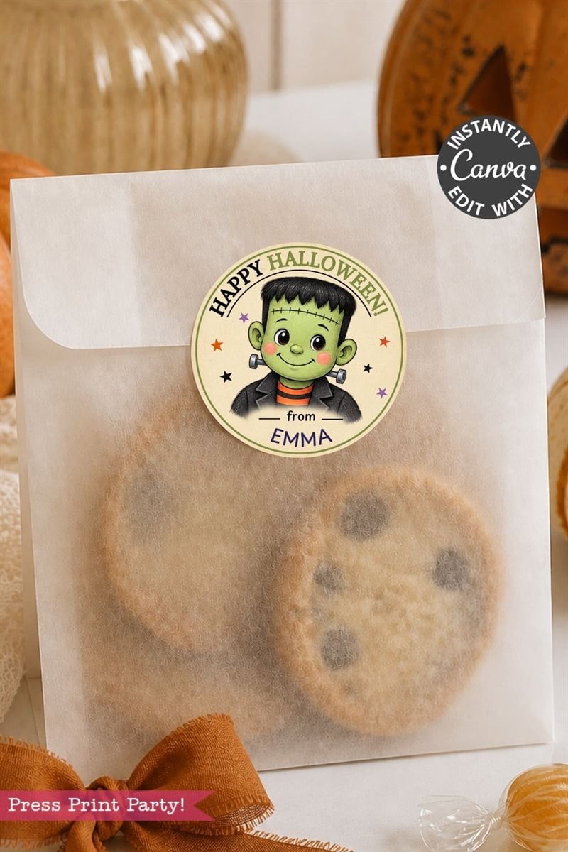 A white treat bag filled with chocolate chip cookies is sealed with a Pink Coquette Ghost Treat Bag Halloween Sticker, featuring a round pink design with a cartoon green Frankenstein and the text “Happy Halloween from EMMA.” Blurred fall decor and a pumpkin appear in the background.
