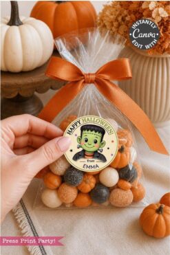 A hand holds a treat bag tied with an orange ribbon, featuring a “Happy Halloween from Emma” sticker with a cartoon Frankenstein and Pink Coquette Ghost Treat Bag Halloween Stickers. Small pumpkins and a cake stand are in the background.
