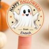A finger holds a Pink Coquette Ghost Treat Bag Halloween Sticker featuring a cute ghost, stars, “Happy Halloween!”, and “from EMMA,” with blurred pumpkins in the background.