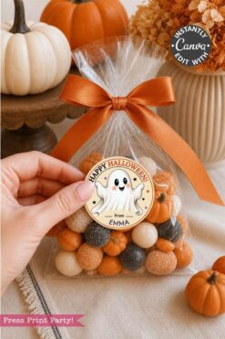 A hand holds a clear bag of orange, white, and black candies tied with an orange ribbon and adorned with a Pink Coquette Ghost Treat Bag Halloween Sticker reading “Happy Halloween from Emma.” Small pumpkins and fall décor are in the background.