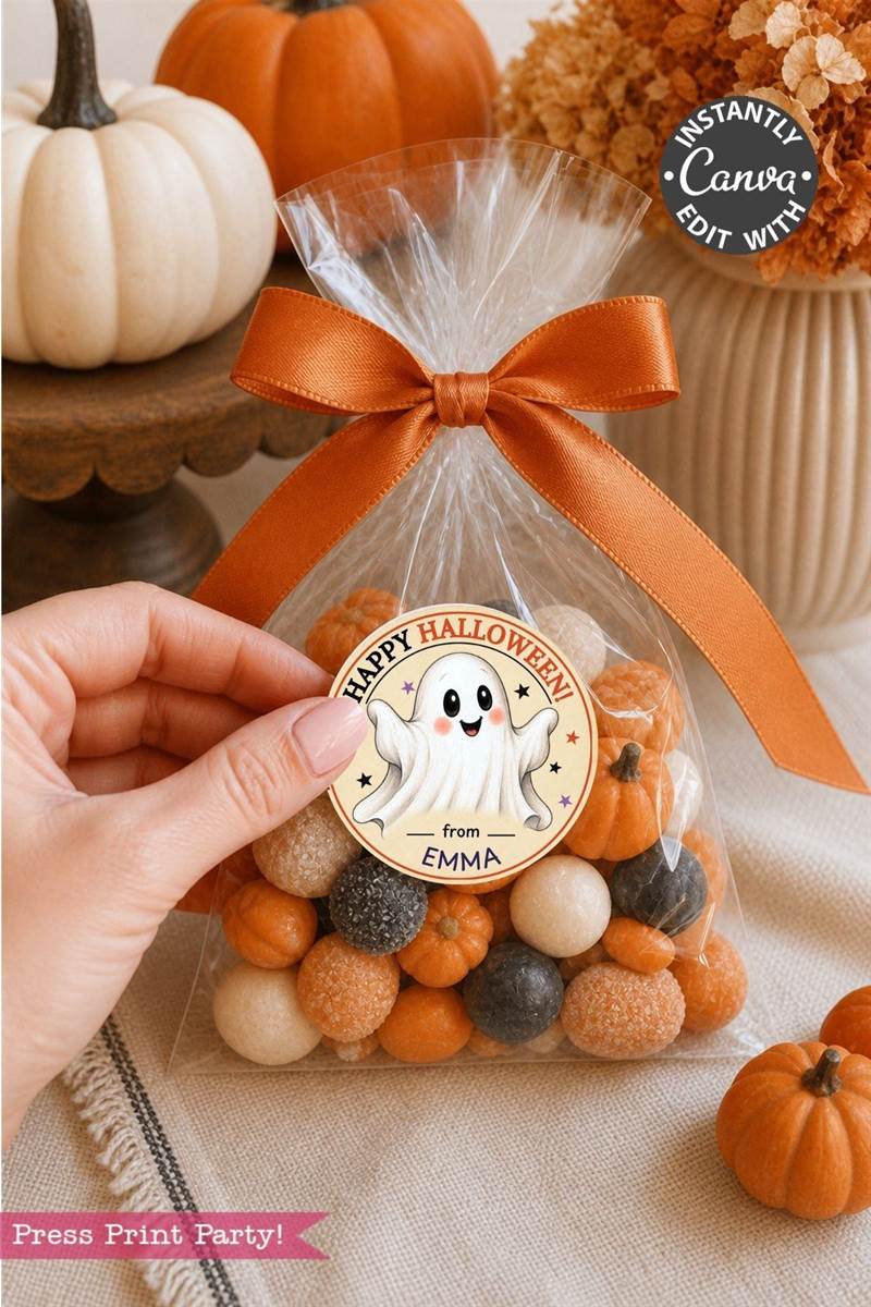 A hand holds a clear bag of orange, white, and black candies tied with an orange ribbon and adorned with a Pink Coquette Ghost Treat Bag Halloween Sticker reading “Happy Halloween from Emma.” Small pumpkins and fall décor are in the background.