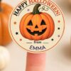 A finger holds a round, pink Coquette Ghost Treat Bag Halloween Sticker featuring a smiling jack-o’-lantern and the text “Happy Halloween! from EMMA” in orange, black, and purple letters. Blurred pumpkins are visible in the background.