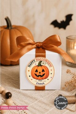 A white gift box with an orange ribbon sits beside a pumpkin and candle. The box features a Halloween label with a smiling jack-o’-lantern, the message “Happy Halloween from Emma,” and Pink Coquette Ghost Treat Bag Halloween Stickers. Dried flowers and wooden beads decorate the scene.