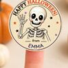 A round Pink Coquette Ghost Treat Bag Halloween Sticker is displayed on a finger, featuring a smiling cartoon skeleton waving with “Happy Halloween! from EMMA” written on it. The cheerful pink coquette Halloween stickers, accompanied by playful pumpkins and a blurred background, create an extra festive vibe.