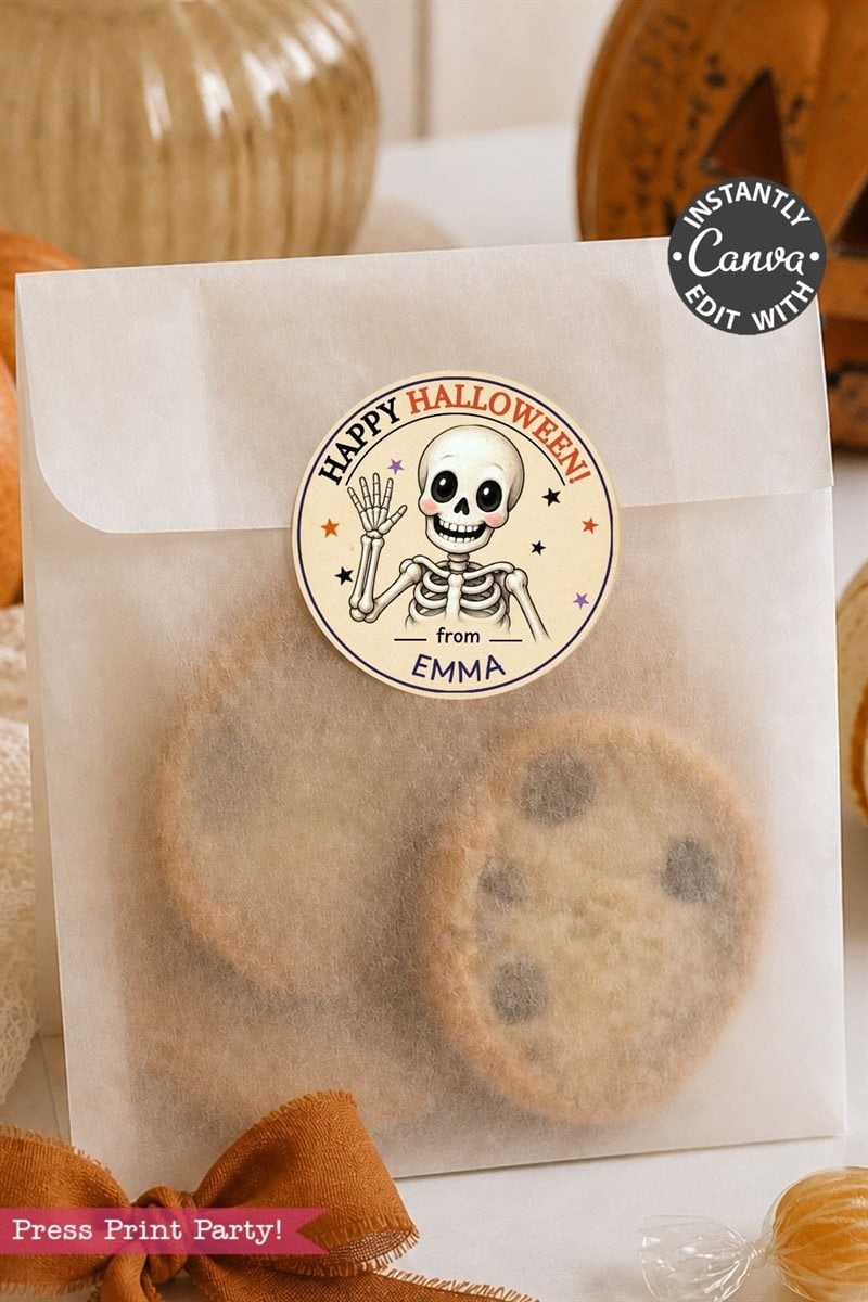 A treat bag filled with cookies is sealed with a Pink Coquette Ghost Treat Bag Halloween Sticker featuring a waving cartoon skeleton, the message “Happy Halloween from Emma,” and small stars, with Halloween decorations visible in the background.
