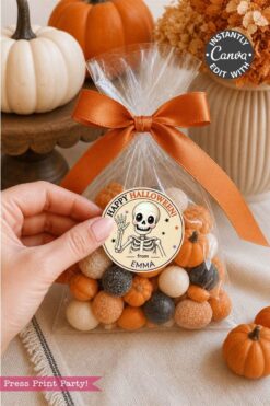 A hand holds a clear treat bag tied with an orange ribbon, filled with orange, white, and black candies. The bag is adorned with a round Halloween label from the Pink Coquette Ghost Treat Bag Halloween Stickers collection, featuring coquette-inspired decorations and a skeleton with “Happy Halloween from Emma.” Small pumpkins decorate the table.