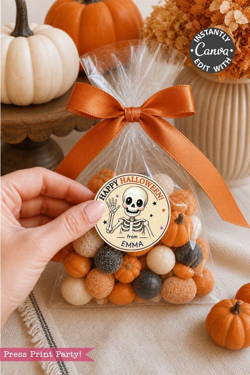 A hand holds a clear treat bag tied with an orange ribbon, filled with orange, white, and black candies. The bag is adorned with a round Halloween label from the Pink Coquette Ghost Treat Bag Halloween Stickers collection, featuring coquette-inspired decorations and a skeleton with “Happy Halloween from Emma.” Small pumpkins decorate the table.