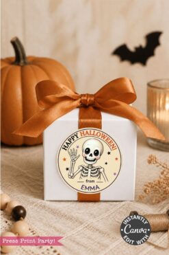 A white gift box with an orange ribbon sits on a table. Adorning the box is a Pink Coquette Ghost Treat Bag Halloween Sticker, featuring a cheery ghost and the message “Happy Halloween from Emma.” In the background, autumnal decor such as a pumpkin and candle complete the festive scene.