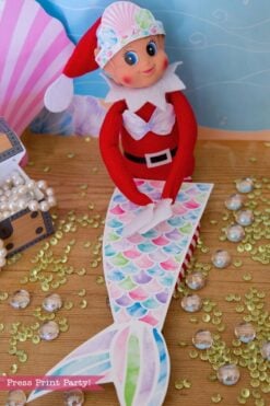 A toy elf wearing a mermaid tail cutout, seashell headband, and shell top sits next to a treasure chest of pearls and beads as part of an Elf Arrival Printable Bundle, Christmas Elf Kit.