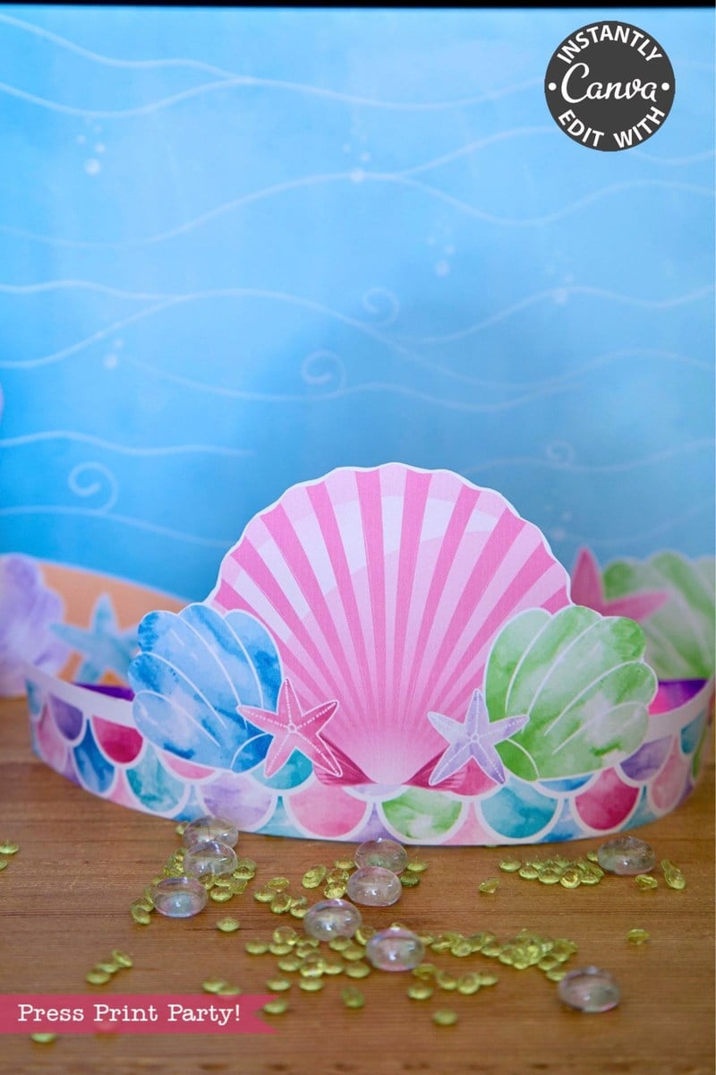 A paper mermaid crown from the Elf Arrival Printable Bundle, Christmas Elf Kit, featuring pink, blue, and green seashell designs and small starfish on a wooden surface with scattered decorative glass beads.