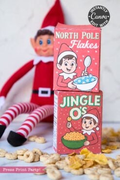 A toy elf sits behind two small, stacked cereal boxes labeled North Pole Flakes and Jingle O’s, surrounded by loose cereal pieces as part of an Elf Arrival Printable Bundle, Christmas Elf Kit.