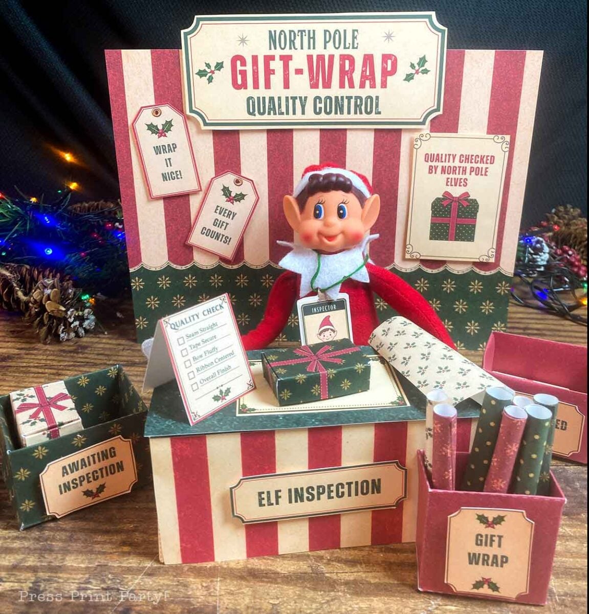 An elf figurine sits at an βElf Inspectionβ station surrounded by boxes of gift wrap and a βNorth Pole Gift-Wrap Quality Controlβ sign.